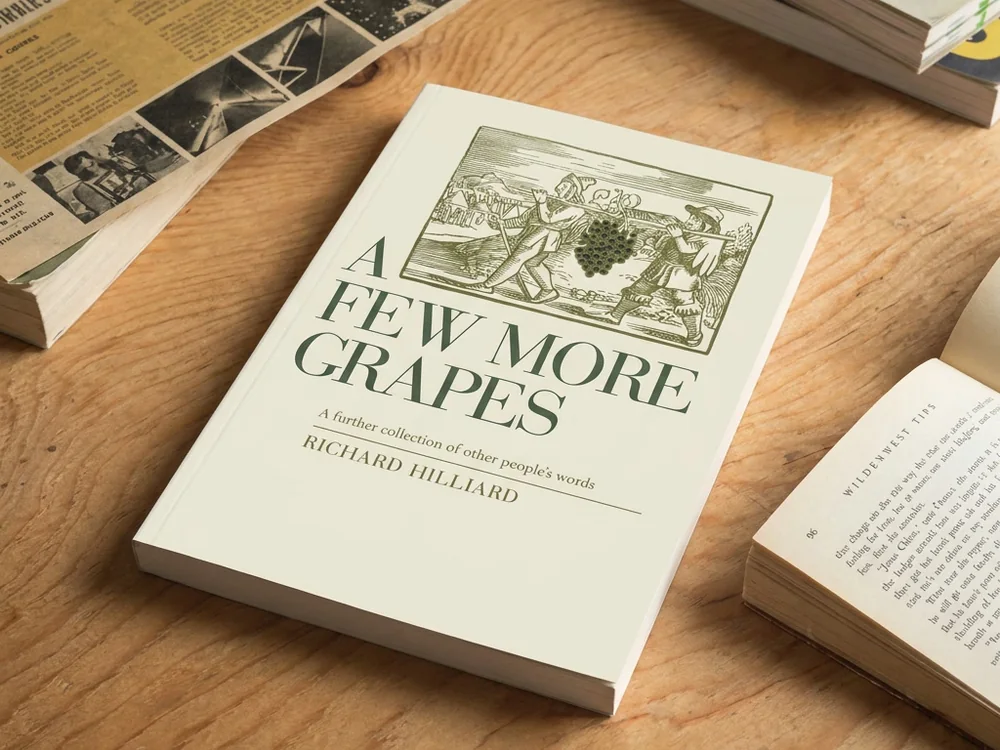 A Few More Grapes by Richard Hilliard, a cream paperback with a green woodcut cover, on a wooden table among books and an open page.